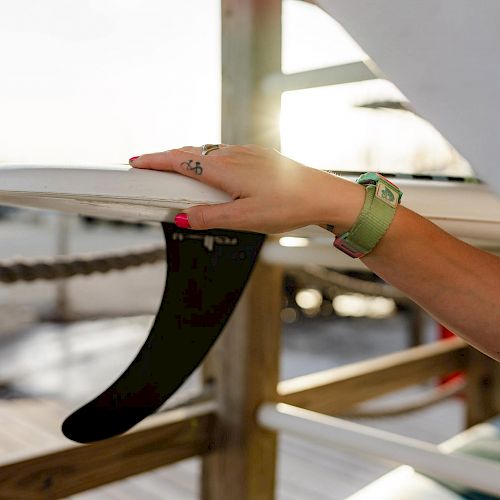 A person's hand with a green watch and red nails is holding the fin of a surfboard, with a beach and wooden railing in the background.