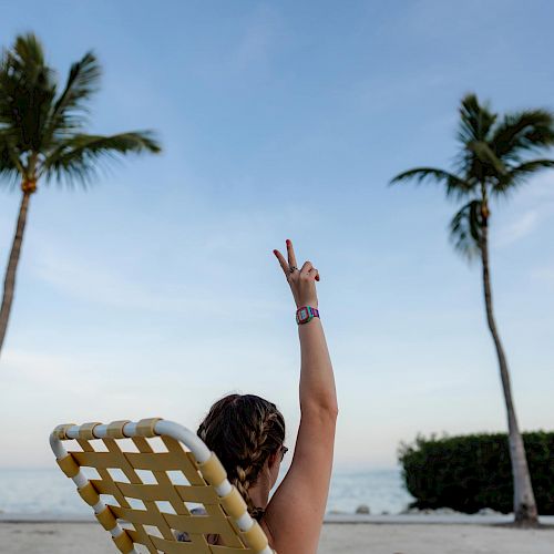 A person sits on a beach chair, raising their arm with a peace sign, surrounded by palm trees and an ocean backdrop, with clear skies.