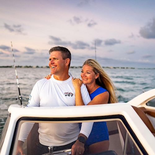 A man and woman are enjoying a boat ride on a body of water during sunset, smiling and standing close to each other, with fishing poles in the background.