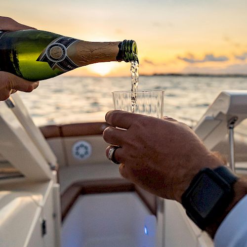 A person pours champagne into a glass on a boat at sunset, with the ocean and a clear sky in the background.