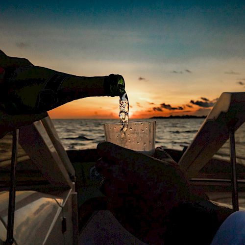 A hand pours a drink into a cup on a boat during sunset over the ocean, with silhouettes in the foreground.