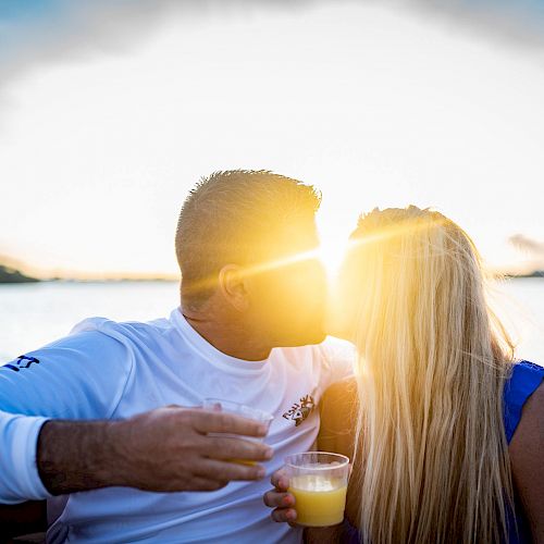 A couple is kissing on a boat during sunset, each holding a drink in their hand.