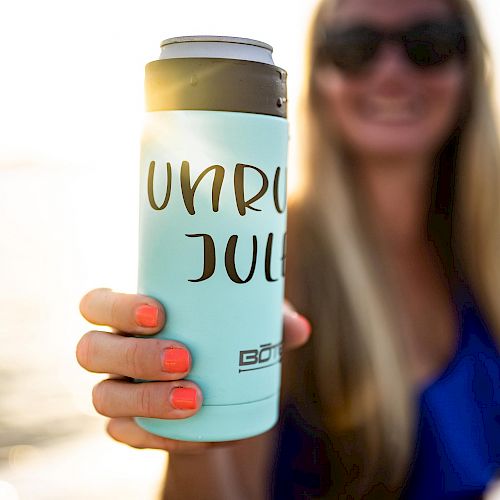 A person with sunglasses holds out a light blue drink holder that says “UNRU JUL” with a can inside. Water and a sunset are in the background.