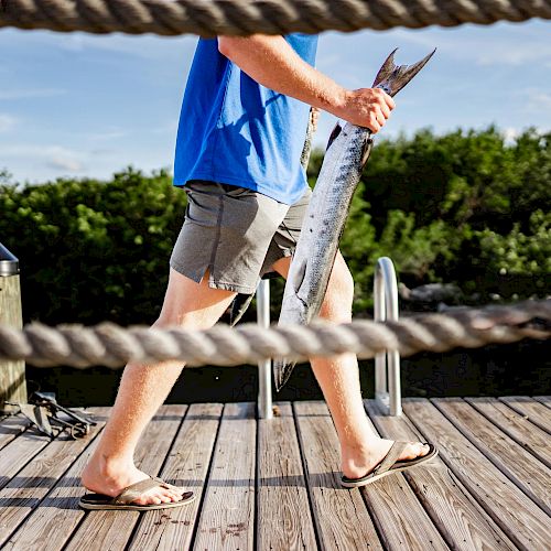 A person in shorts and flip-flops walks on a dock holding a large fish with green foliage and water in the background.