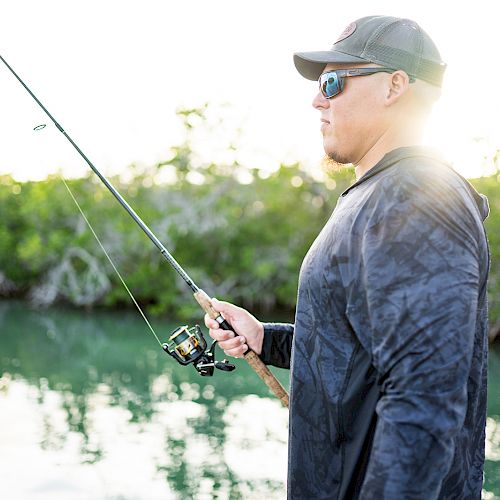 A person is fishing near water, holding a fishing rod, wearing a cap and sunglasses, with a backdrop of greenery and sunlight.