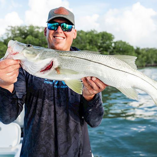 A person wearing sunglasses and a cap is holding a large fish on a boat, with a water and green foliage background.