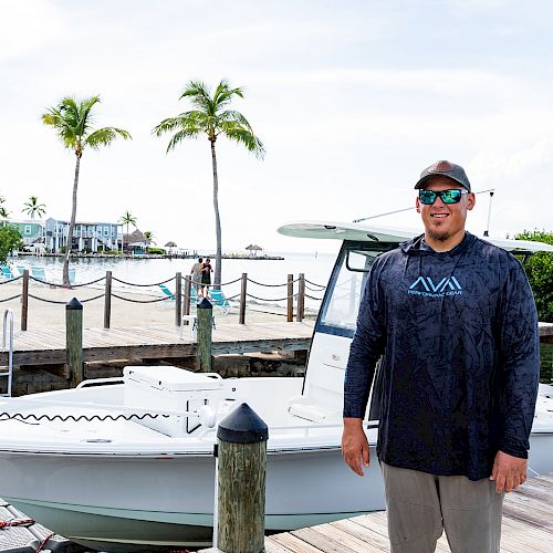 A person standing on a dock beside a white boat, with palm trees and buildings in the background on a sunny day.