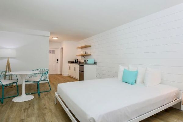 A modern, minimalist bedroom with a large bed, turquoise-accented chairs and cushions, a small dining area, and a kitchenette in a bright, white room.