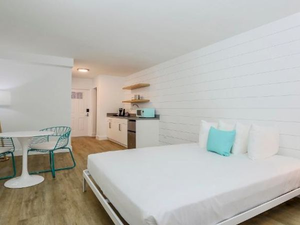 A modern, minimalist bedroom with a large bed, turquoise-accented chairs and cushions, a small dining area, and a kitchenette in a bright, white room.