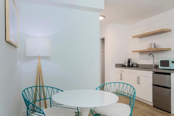 This image shows a modern kitchen with a small round table, two chairs, a floor lamp, open shelves, a sink, a microwave, and a mini fridge.