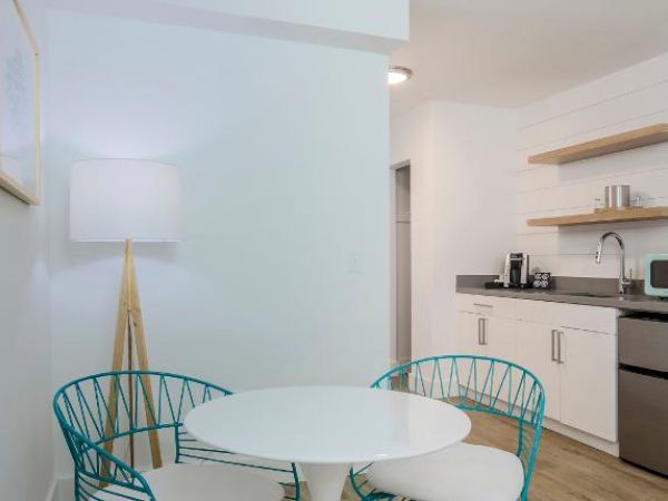 This image shows a modern kitchen with a small round table, two chairs, a floor lamp, open shelves, a sink, a microwave, and a mini fridge.