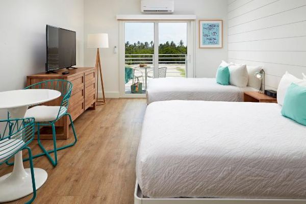A bright, modern hotel room with two beds, a flat-screen TV, a dresser, a small table with chairs, and a balcony overlooking greenery.