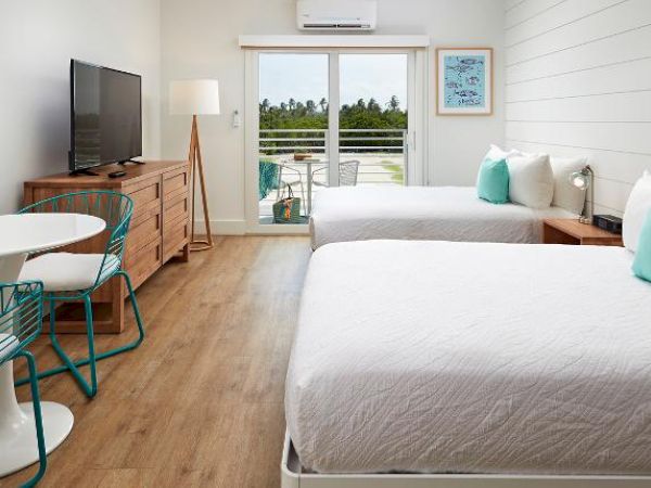 A bright, modern hotel room with two beds, a flat-screen TV, a dresser, a small table with chairs, and a balcony overlooking greenery.