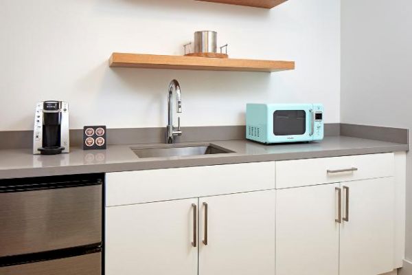 The image shows a modern kitchenette with a sink, a microwave, a coffee maker, a mini-fridge, and open shelves.