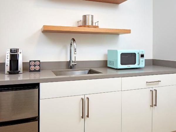 The image shows a modern kitchenette with a sink, a microwave, a coffee maker, a mini-fridge, and open shelves.