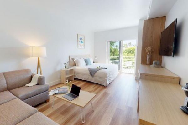 A modern, well-lit studio apartment features a bed, couch, coffee table, lamp, TV, desk, and a coffee maker, with a balcony overlooking greenery.