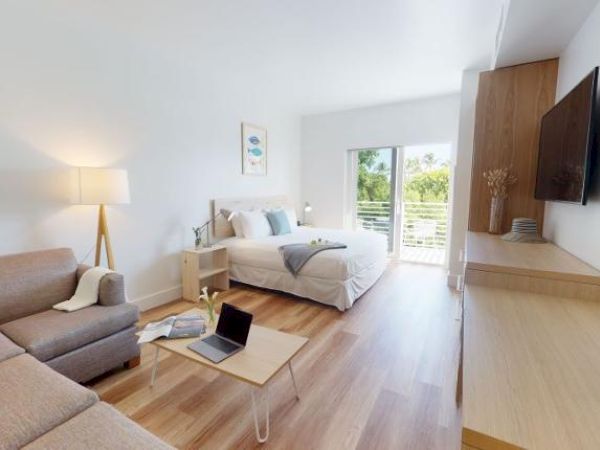 A modern, well-lit studio apartment features a bed, couch, coffee table, lamp, TV, desk, and a coffee maker, with a balcony overlooking greenery.