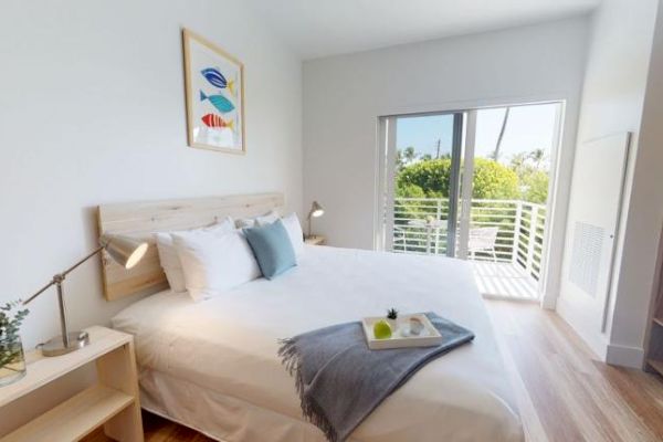 A bright, modern bedroom with a large bed, wall art, and patio doors opening to a balcony with greenery. Accessories include a tray on the bed.