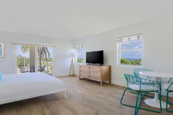 A bright, airy room with a bed, TV on a wooden stand, round table with chairs, and a balcony with a view of palm trees and greenery.