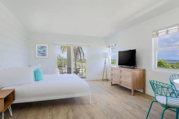 A bright and airy bedroom features a bed, side tables, TV, dresser, and a small dining table. Sliding doors lead to a balcony with a view.