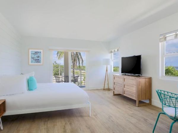 A bright and airy bedroom features a bed, side tables, TV, dresser, and a small dining table. Sliding doors lead to a balcony with a view.