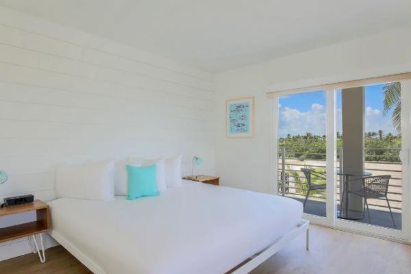 A modern, bright bedroom with a large bed, nightstands, wall art, and sliding doors opening to a balcony with a scenic view.