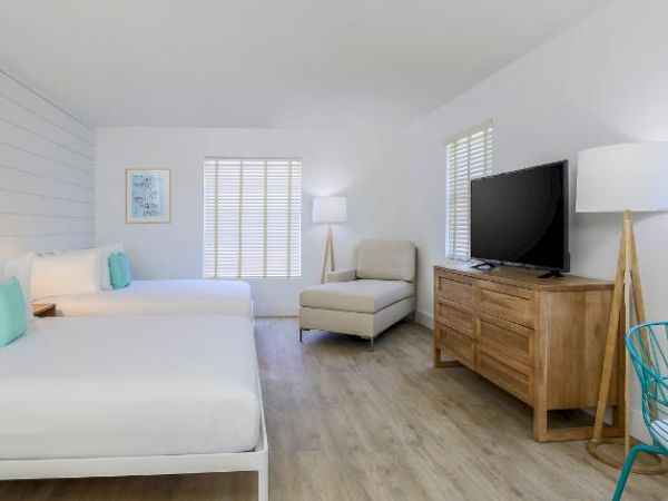This image shows a bright, modern hotel room with two beds, a TV on a wooden dresser, an armchair, a floor lamp, and a small table with a chair.