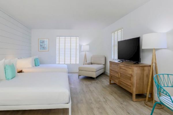 The image shows a modern, bright hotel room with two beds, a wooden dresser, a large TV, a chair, and a lamp. The room has wooden floors.