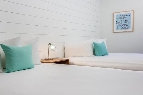 A clean, minimalistic bedroom with two beds, teal pillows, a small bedside table with a lamp, and wall art featuring fish.