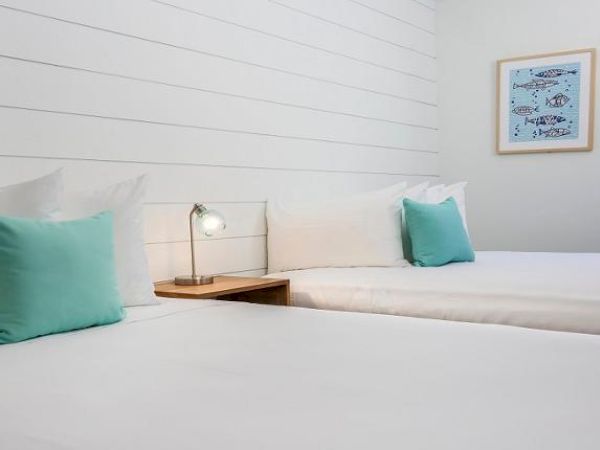 A clean, minimalistic bedroom with two beds, teal pillows, a small bedside table with a lamp, and wall art featuring fish.