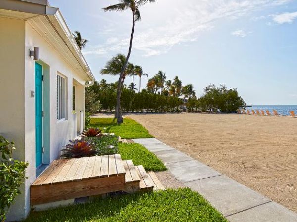 A pristine beach with palm trees, sun loungers, and a bright beach house with a turquoise door. The ocean is in the background under a clear sky.