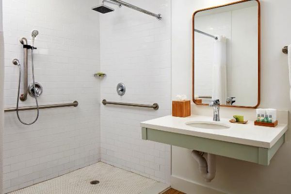 The image shows a modern bathroom with a walk-in shower, grab bars, a shower bench, a sink with a mirror, and toiletries on the counter.