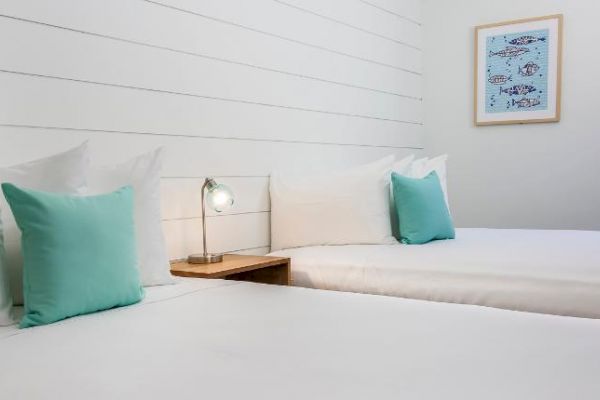 The image shows a clean, minimalist bedroom with two beds, each with a green pillow, a small lamp on a wooden nightstand, and a nautical-themed picture.