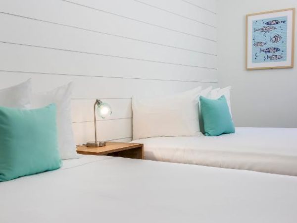 The image shows a clean, minimalist bedroom with two beds, each with a green pillow, a small lamp on a wooden nightstand, and a nautical-themed picture.