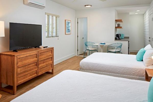 The image shows a modern, well-lit hotel room with two beds, a TV on a wooden dresser, a small dining area, and a kitchenette.