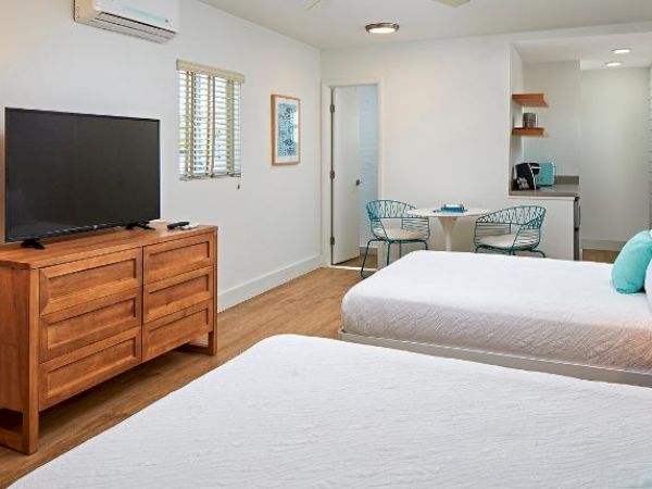 The image shows a modern, well-lit hotel room with two beds, a TV on a wooden dresser, a small dining area, and a kitchenette.