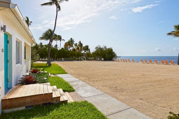 A beachfront scene shows a house with a turquoise door, sand, palm trees, and orange beach chairs facing the sea under a blue sky.