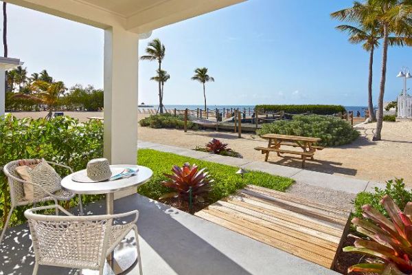 A beachside patio with chairs, a table, and plants, overlooking a path, picnic table, and palm trees leading to the ocean under a clear blue sky.