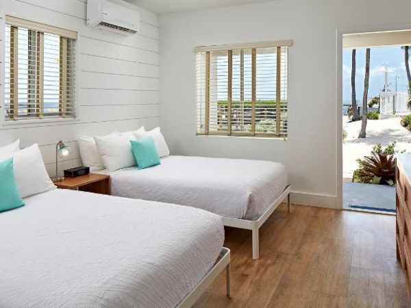 A bright bedroom features two double beds with white linens and turquoise pillows, wooden floor, TV, and a door showing a view of the outdoors.