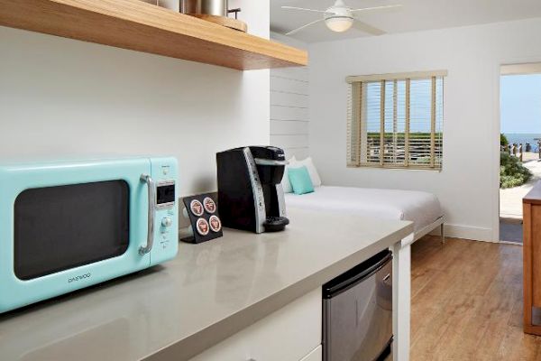 A cozy room with a light blue microwave, a coffee maker, a bed, and a view of the ocean through a door. The room has wooden floors and modern decor.