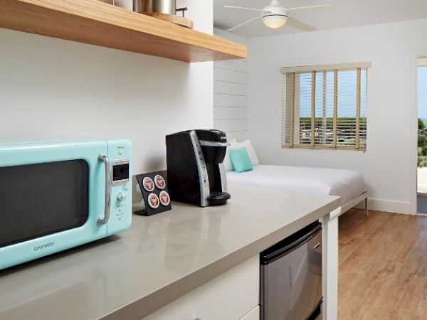 A cozy room with a light blue microwave, a coffee maker, a bed, and a view of the ocean through a door. The room has wooden floors and modern decor.