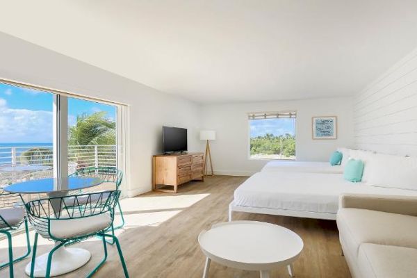 A bright, modern room with ocean views, featuring a bed, sofa, TV, and dining area with a table and chairs, and large windows for natural light.
