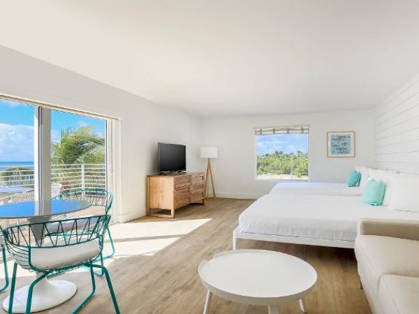 A bright, modern room with ocean views, featuring a bed, sofa, TV, and dining area with a table and chairs, and large windows for natural light.