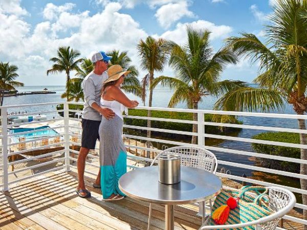 A couple stands on a balcony overlooking a tropical landscape with palm trees, a pool, and a body of water. On the table is a colorful bag.