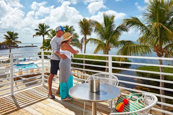 A couple stands on a balcony overlooking a tropical seaside view with palm trees, a pool, and clear skies. A table and chairs are in the foreground.