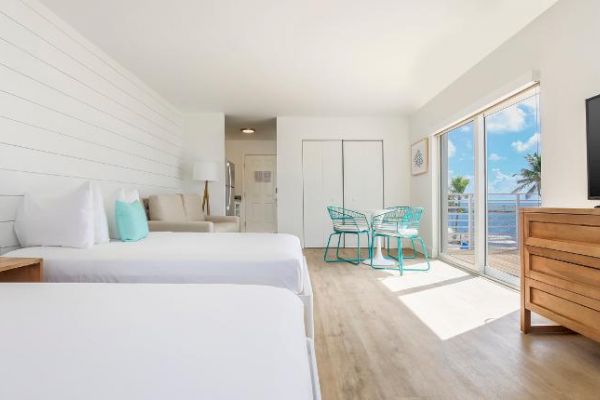 A bright, modern hotel room features two beds, a TV, a small dining table, and a great view of the beach through large sliding glass doors.