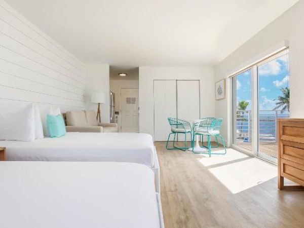 A bright, modern hotel room features two beds, a TV, a small dining table, and a great view of the beach through large sliding glass doors.