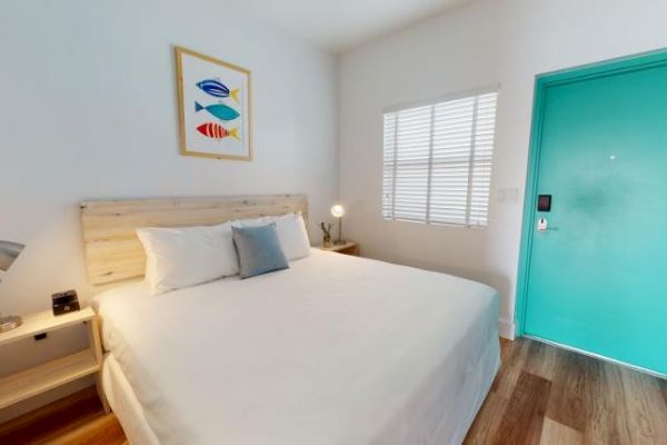 A tidy bedroom features a queen-sized bed, two small nightstands with lamps, a framed fish artwork, a window with blinds, and a turquoise door.