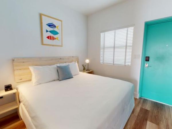 A tidy bedroom features a queen-sized bed, two small nightstands with lamps, a framed fish artwork, a window with blinds, and a turquoise door.