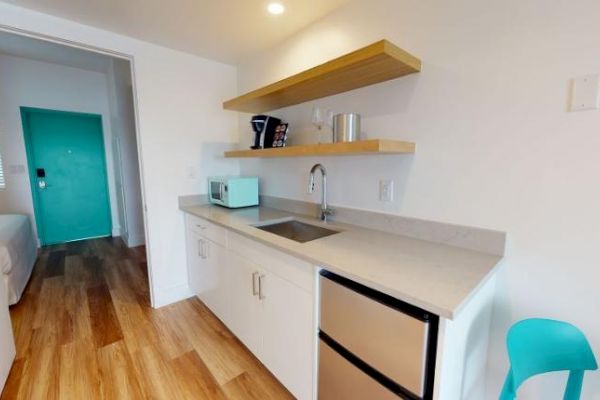 This image depicts a small kitchenette with a sink, a mini fridge, open shelves, a microwave, and a teal chair, leading to a room with a turquoise door.